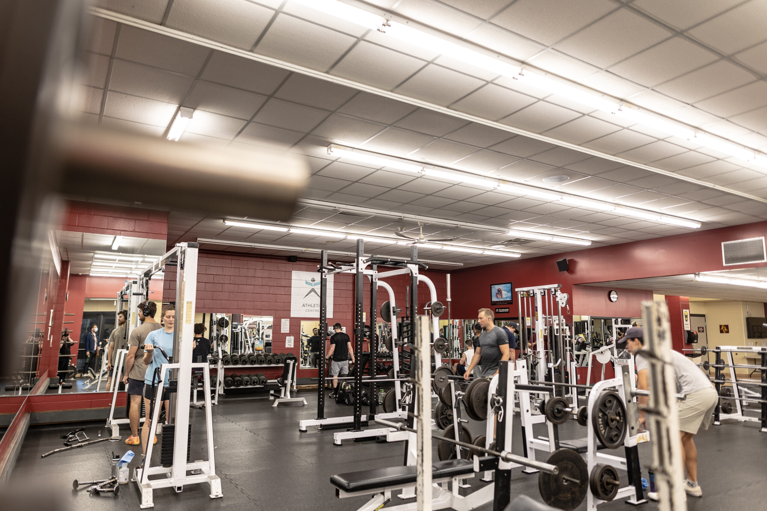 The Cambrian Athletics Centre bustles with activity, as several people engage with weightlifting equipment, mirrors reflecting their determination under the glow of fluorescent lighting.