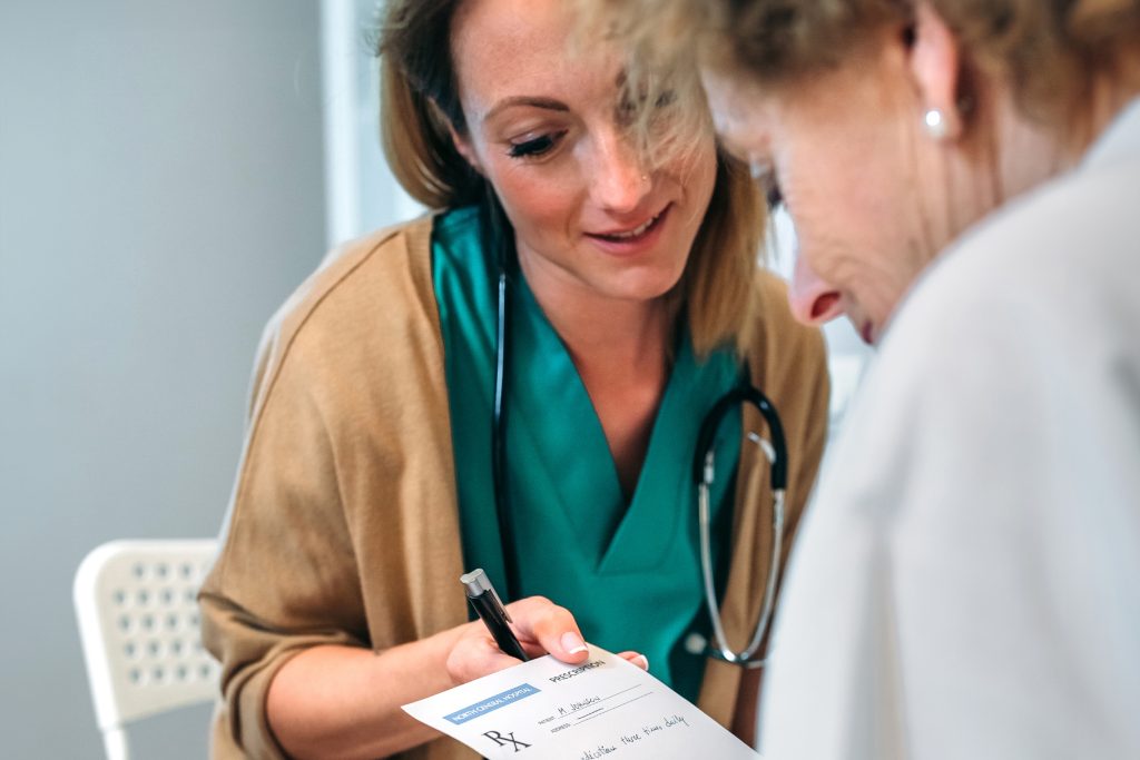 A medical professional in a teal uniform discusses a prescription with an older adult, pointing to the document while holding a pen.