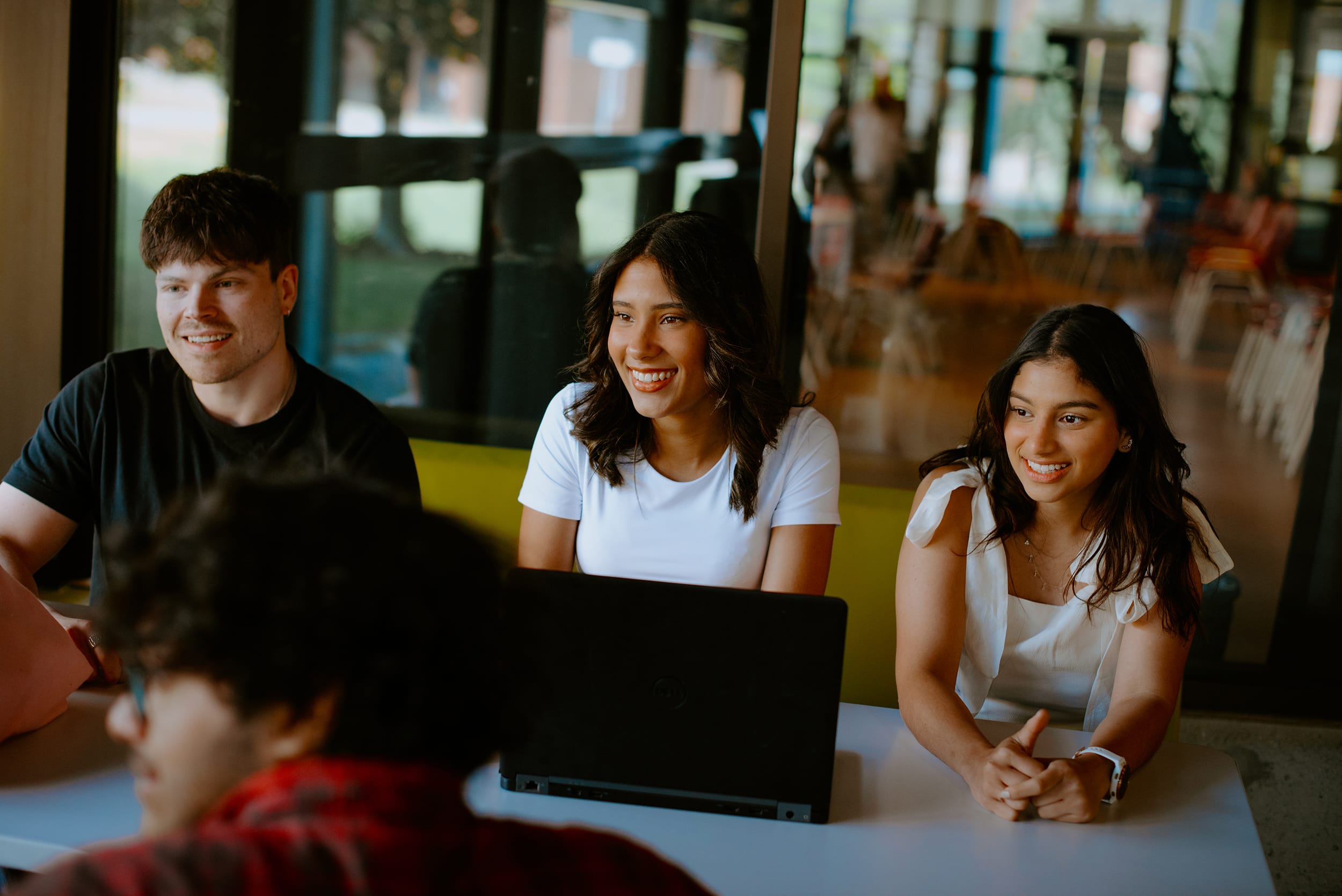 Four students sit around a table with laptops, engaged in conversation. They are indoors, near large windows with a view of the outside.