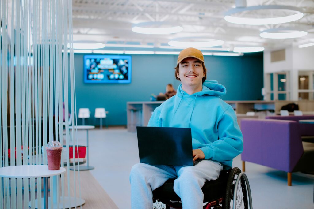 Student with a laptop, wearing a blue hoodie and cap, smiles in a modern lounge area.