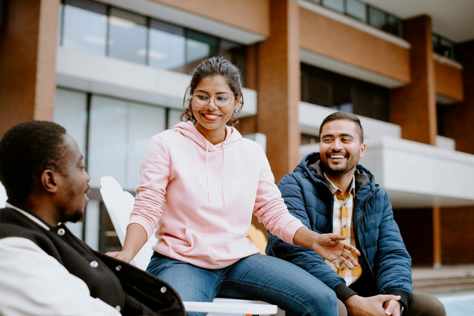 Three student sit and chat outdoors in front of a building. One person wears a pink hoodie, another a blue jacket, and the third a black jacket.