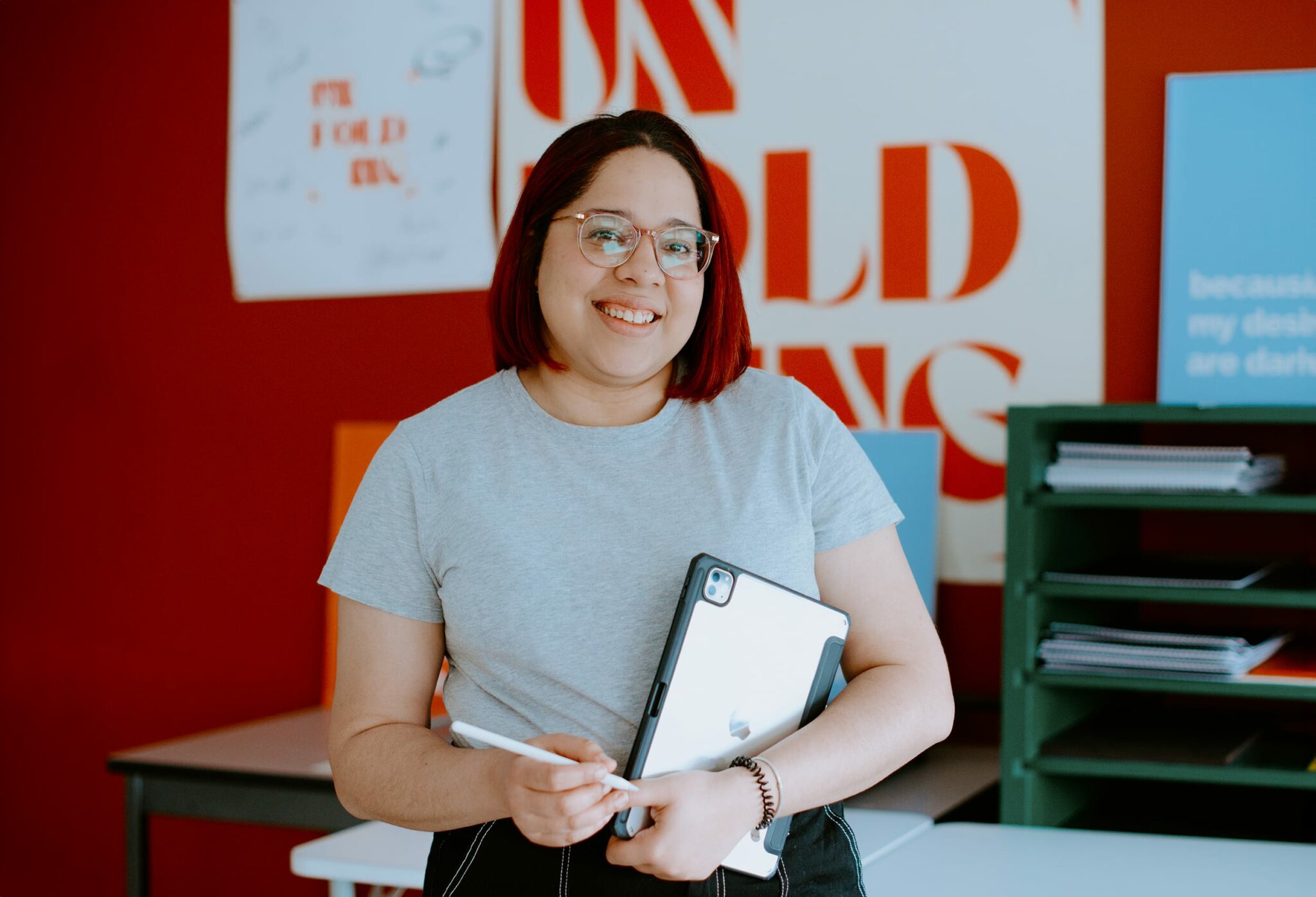 A person with glasses and short red hair, wearing a gray shirt, stands smiling while holding a tablet and stylus. Background includes shelves with papers and red wall with typography.