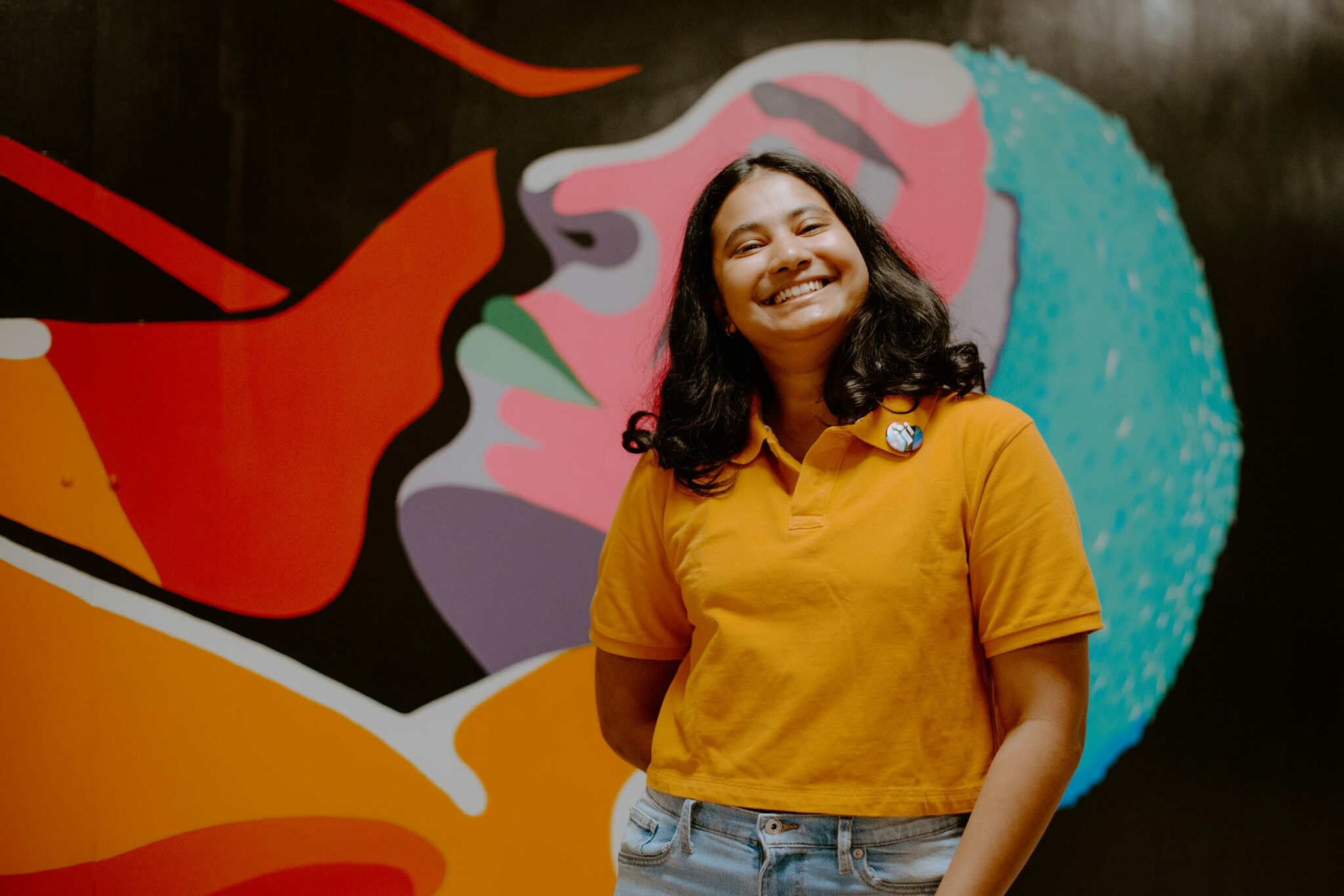 Student in a yellow shirt smiling in front of a colorful abstract mural.