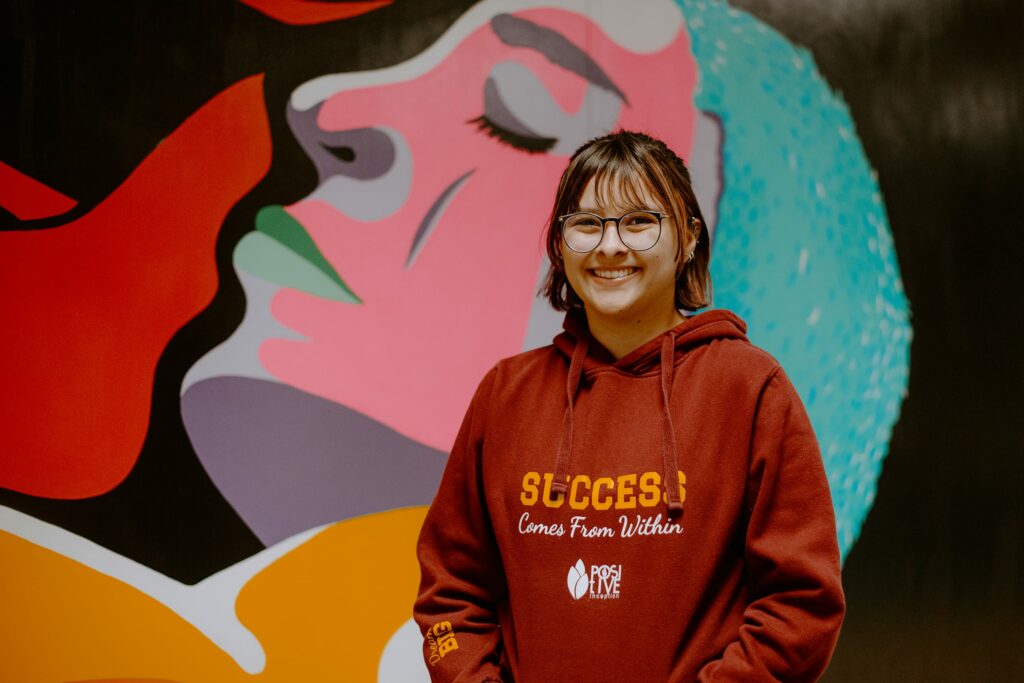 A student in glasses and a maroon hoodie stands smiling in front of a colorful mural featuring abstract faces.