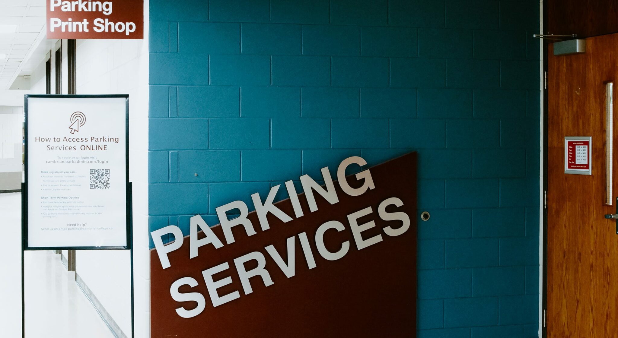 Signage for "Parking Services" and "Parking Print Shop" on a blue wall with a digital access poster within Cambrian College.
