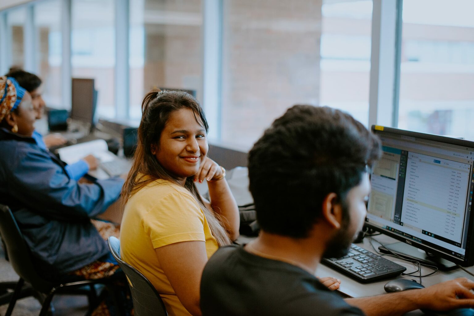 A group of students working at computer stations in a bright room. A student in a yellow shirt smiles at the camera.
