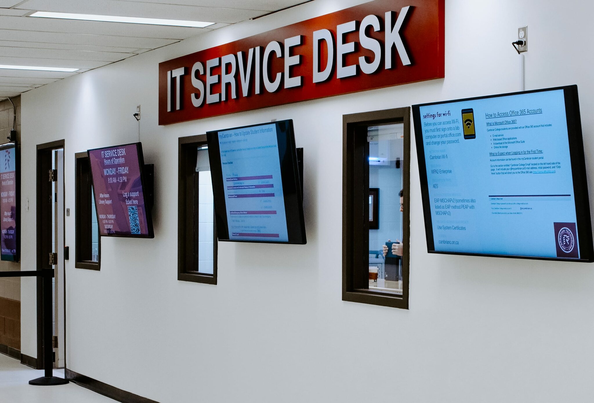 Hallway with Cambrian College's IT Service Desk sign above multiple screens displaying information on a white wall.