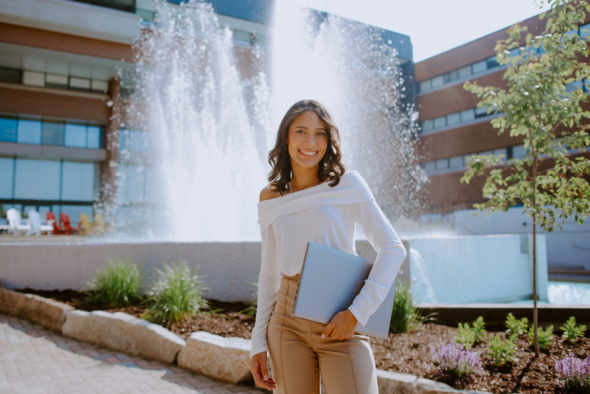 Student in a white top and beige pants stands smiling, holding a laptop in front of a fountain with a modern building in the background.