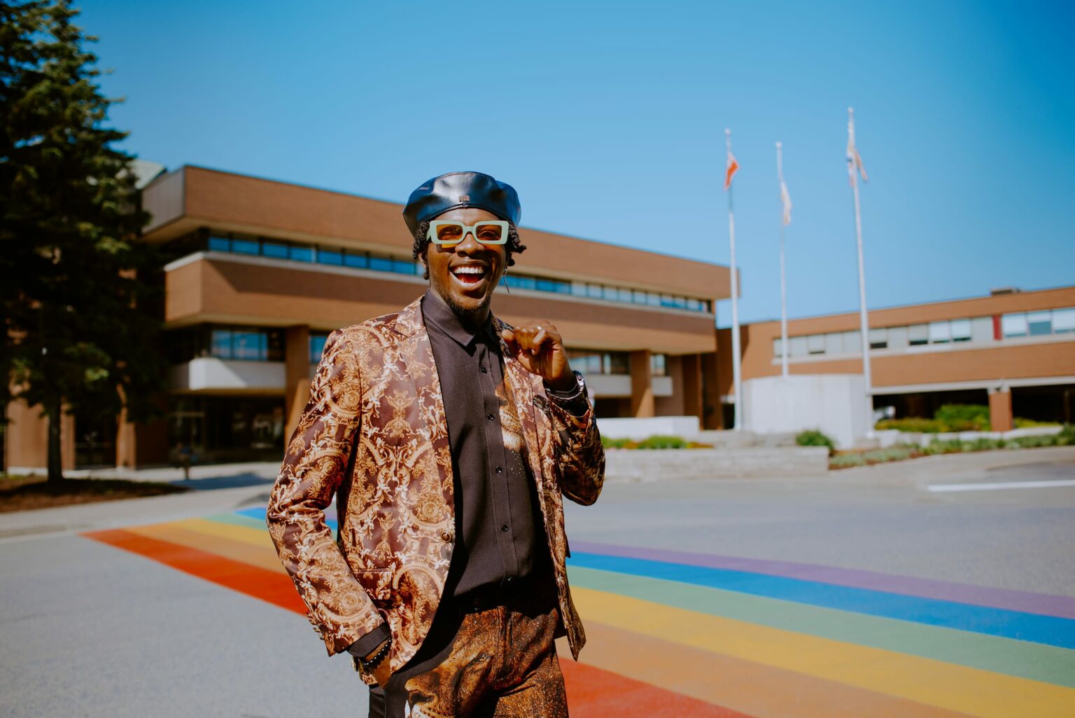 Student in a patterned suit and beret stands on a rainbow crosswalk in front of a building with flags, smiling under a clear blue sky.