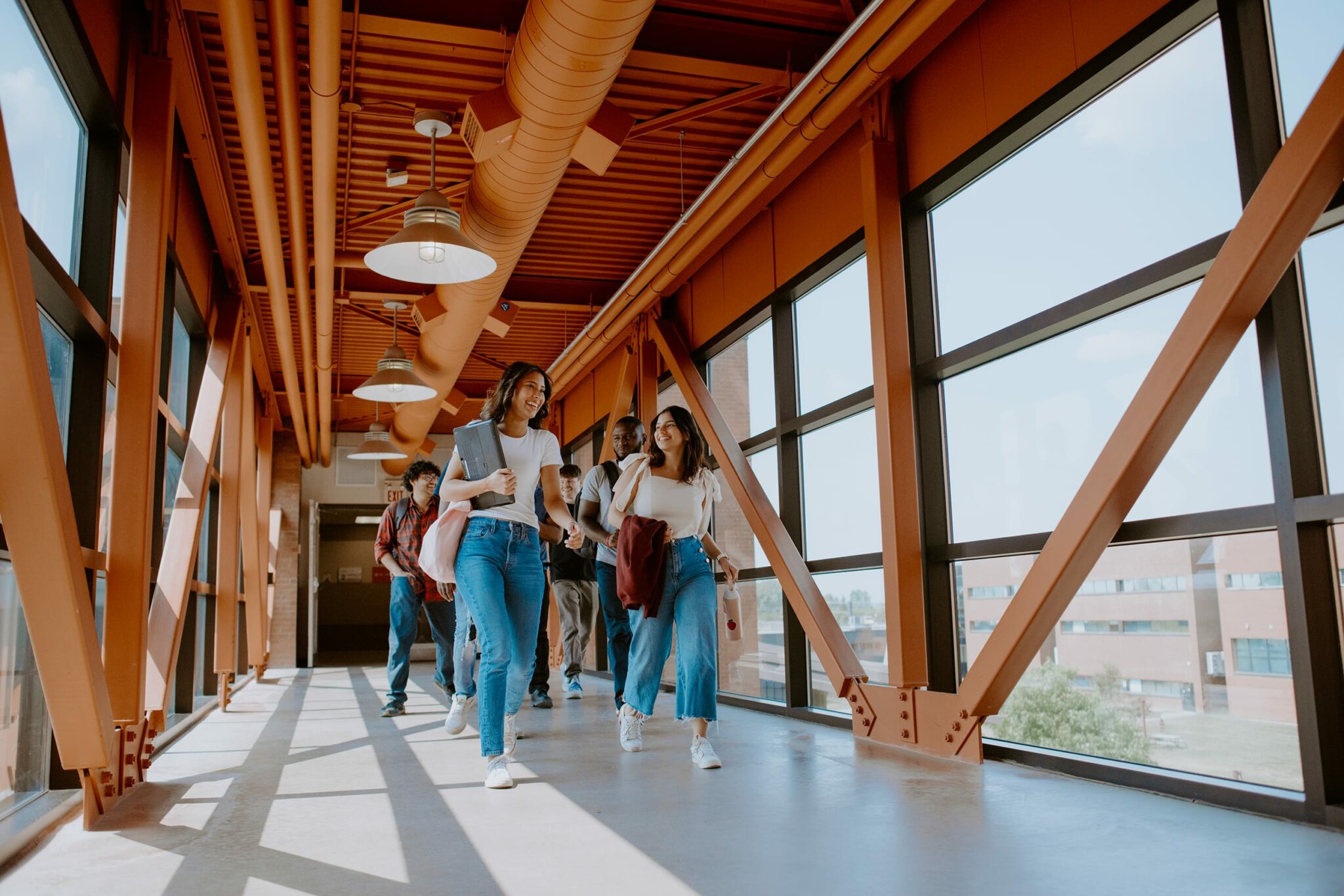 A group of students walk along a glass-walled hallway with orange beams. They carry backpacks and books.