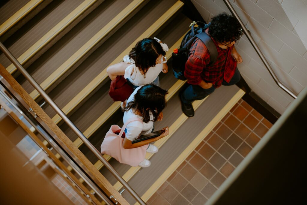 Three students walk down a staircase in a building. They are carrying backpacks and wearing casual clothing.