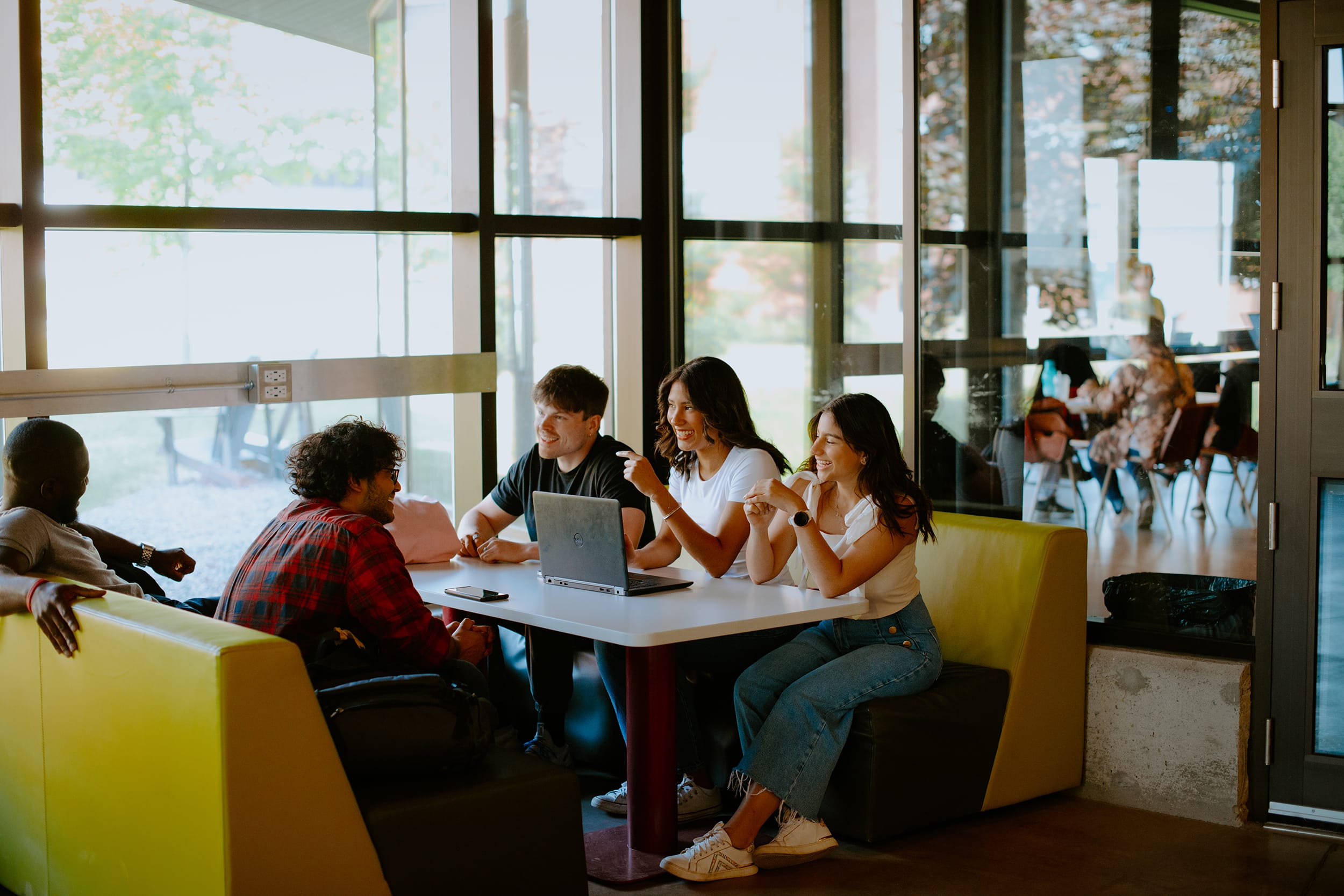 A group of five students sit around a table with a laptop, talking and smiling, inside a room with large windows.