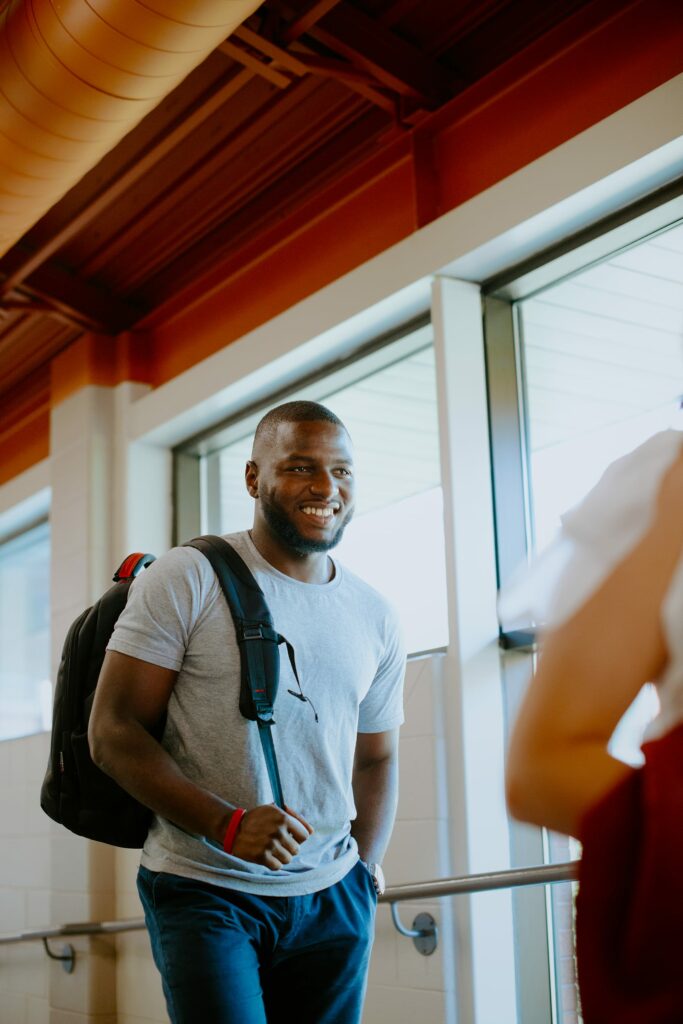 A student wearing a gray t-shirt and carrying a backpack walks indoors, smiling. He is near a large window in a brightly lit space with red structural beams overhead.