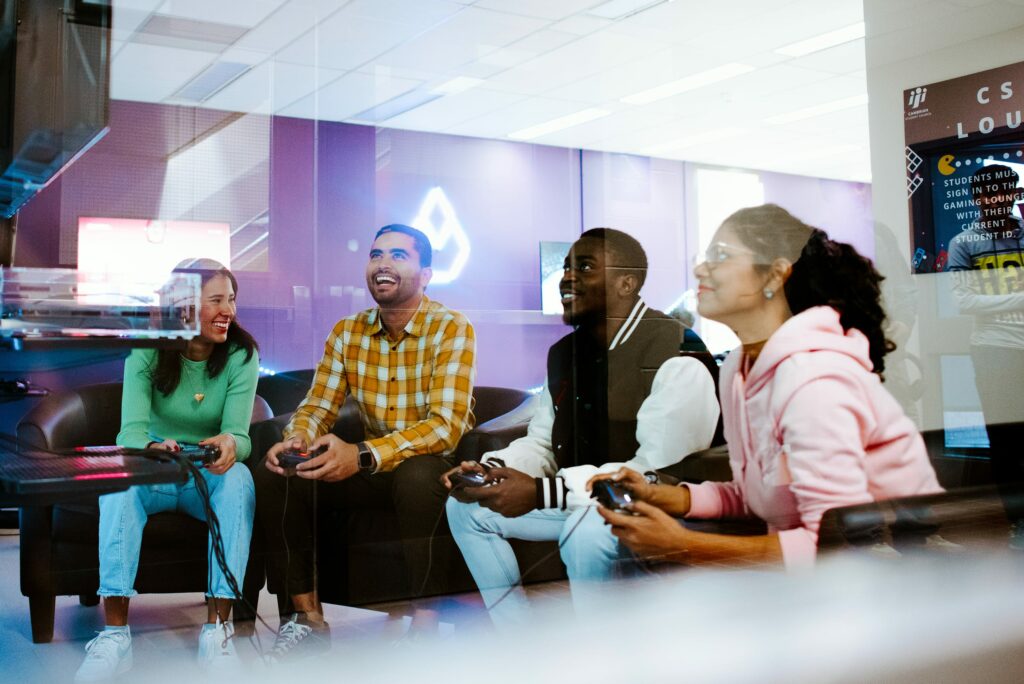 Four students sitting on couches are playing video games, smiling and holding controllers in a brightly lit room.