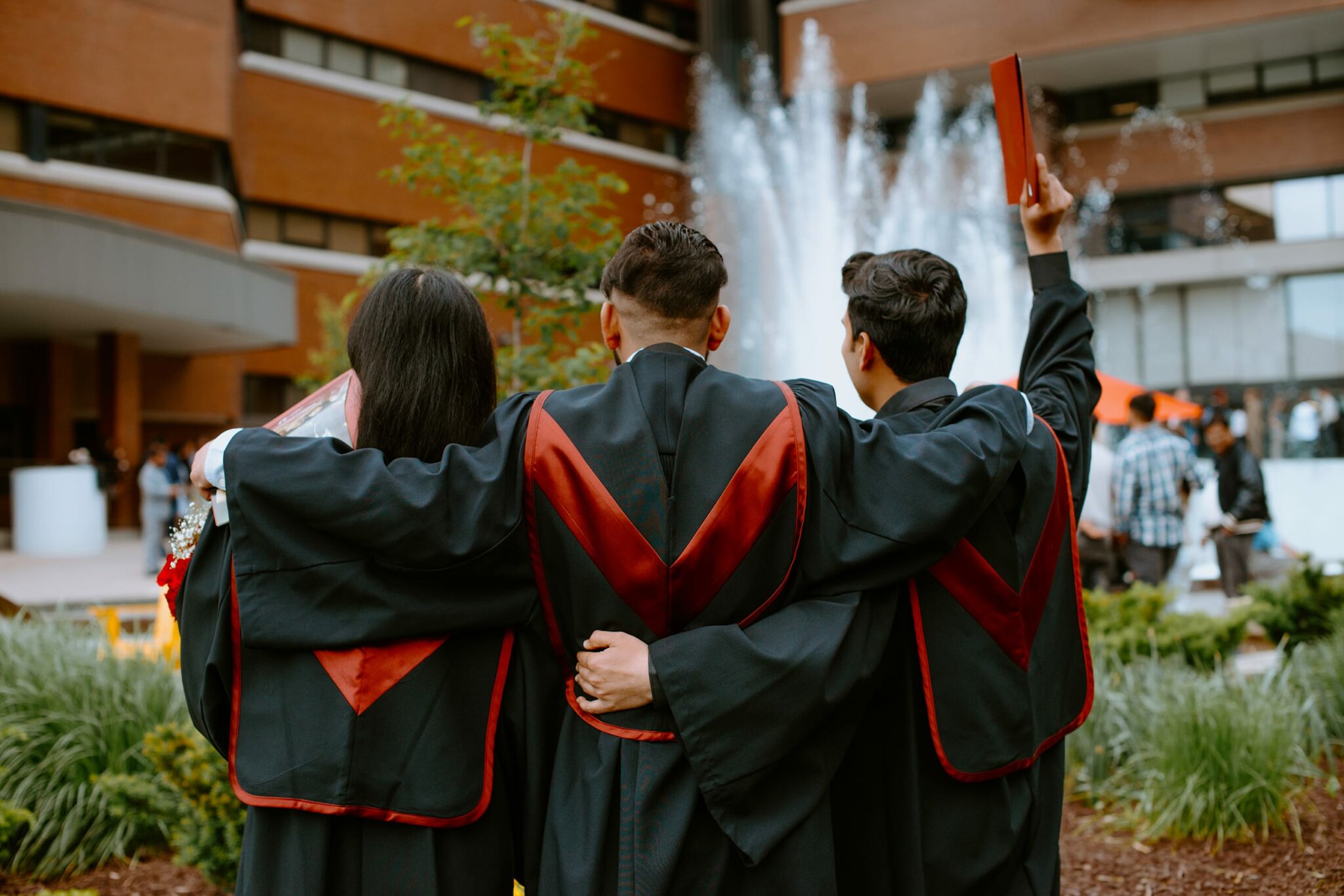 Three graduates in caps and gowns stand with arms around each other, facing a fountain and holding diplomas.