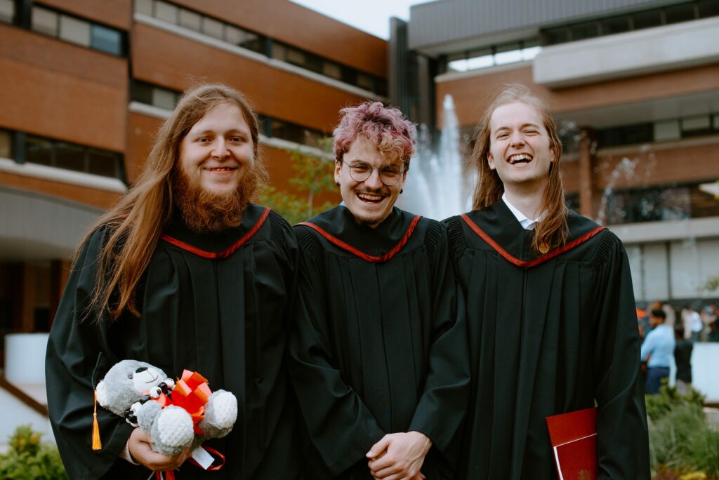 Three graduates in black gowns smiling outdoors, one holding a teddy bear, with a building and fountain in the background.