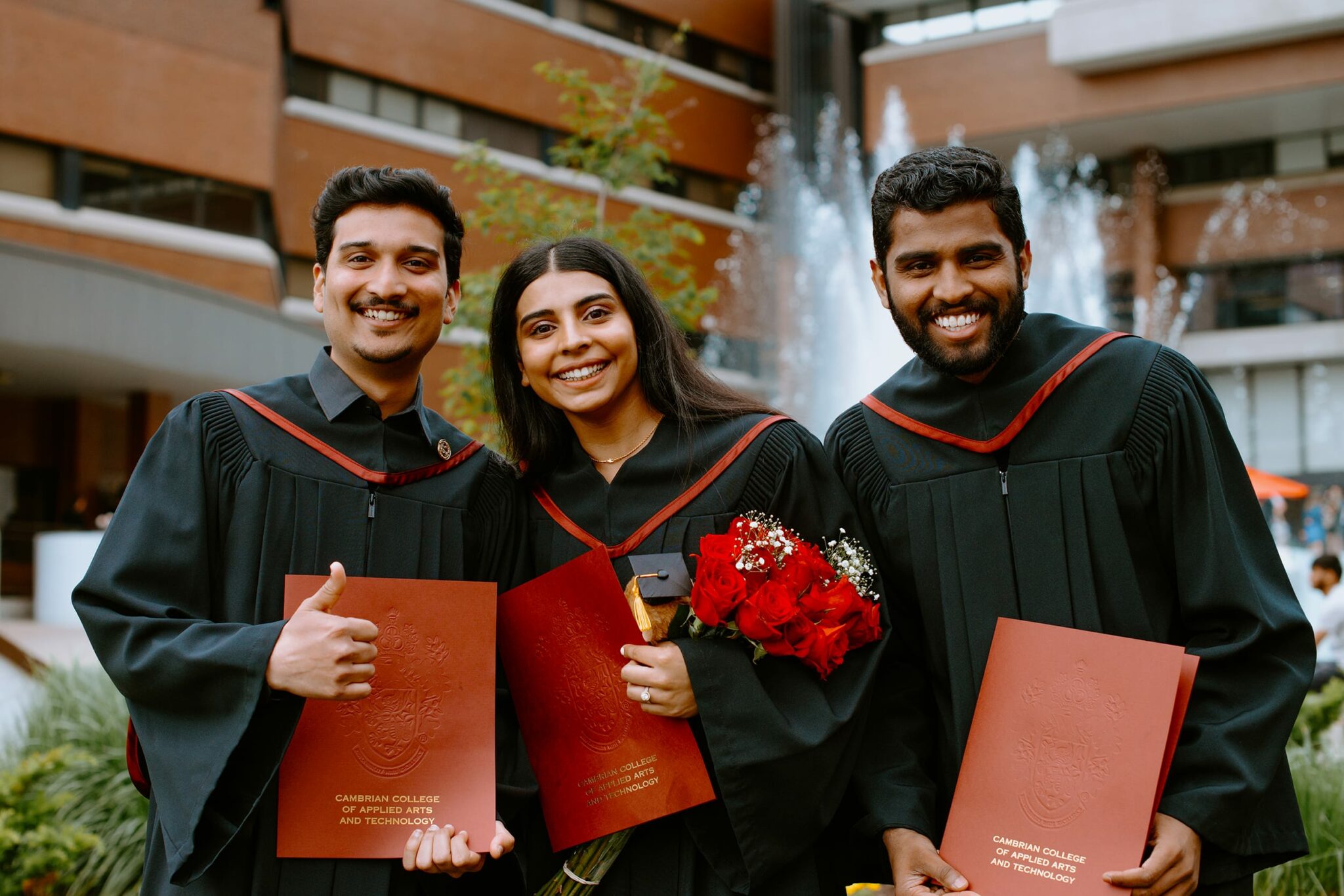 Three graduates in black gowns pose with diplomas and a bouquet of red roses, smiling in front of a fountain at a college campus.