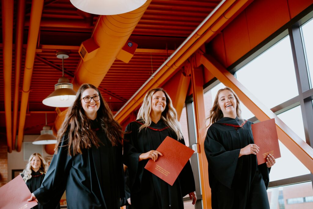Group of graduates in caps and gowns holding diplomas, walking indoors under red beams and lighting.