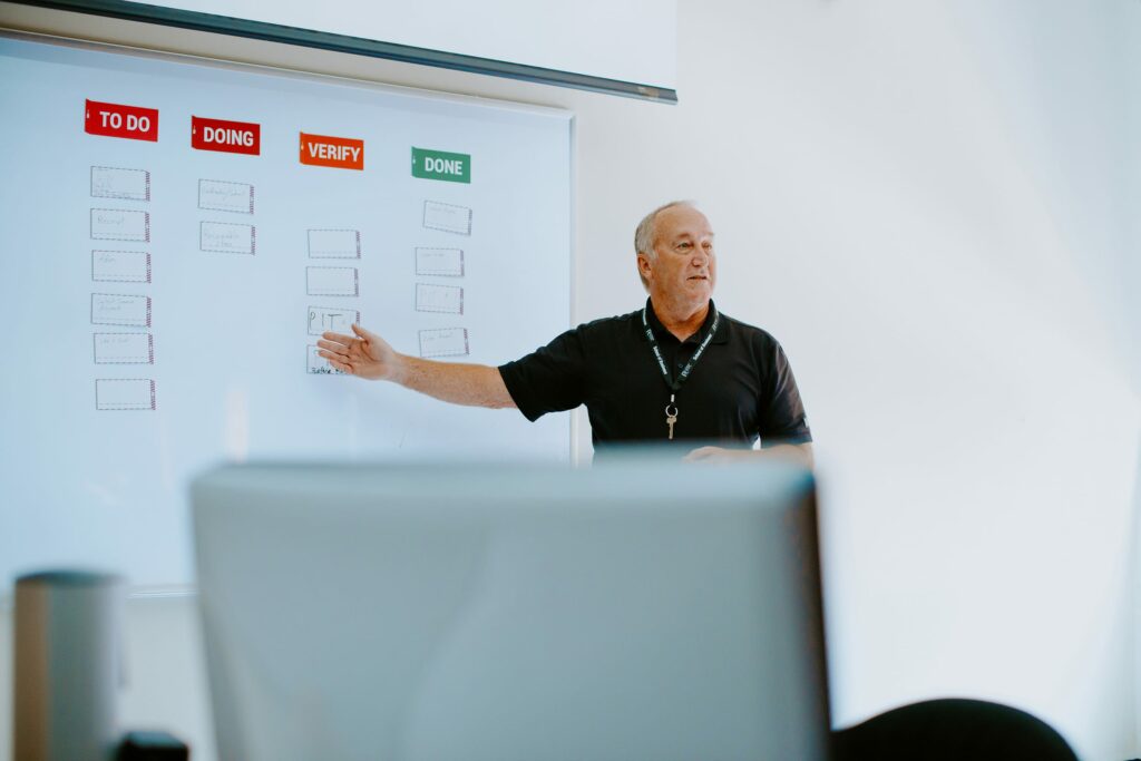 A staff member in a black shirt stands in front of a whiteboard with columns labeled "TO DO," "DOING," "VERIFY," and "DONE," pointing to a task. A computer monitor is in the foreground.
