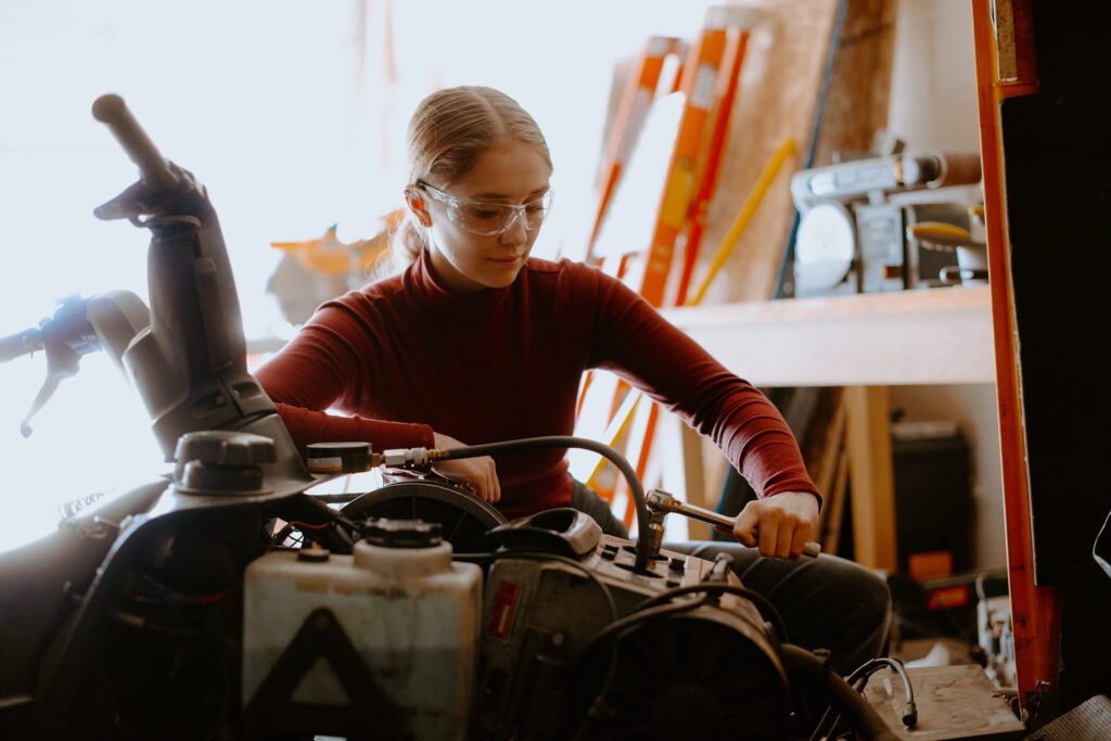 A student wearing goggles is working on a machine in a workshop. Various tools and equipment are visible in the background.