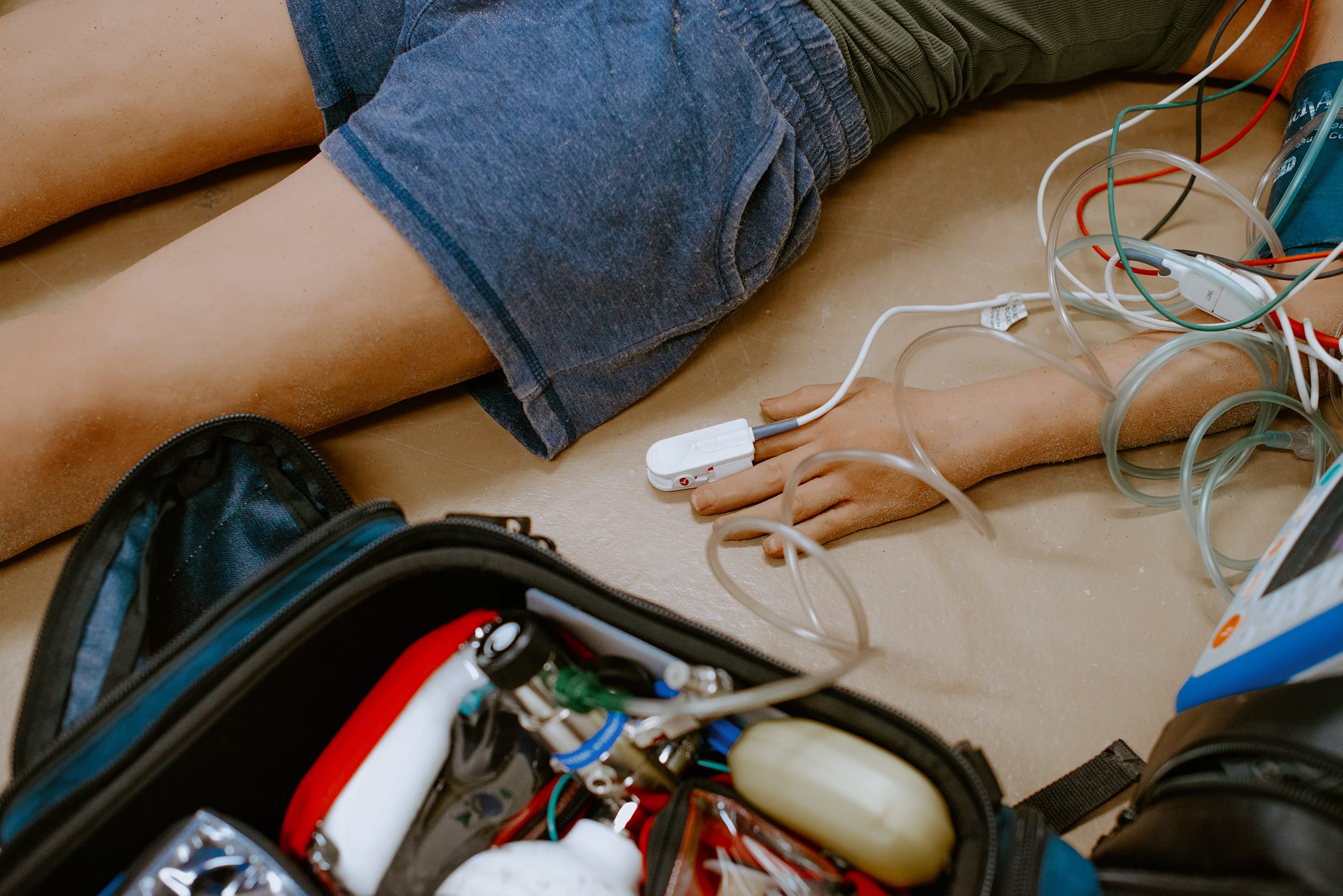 Person lying on the ground with medical equipment, including a pulse oximeter on their finger, surrounded by medical supplies.