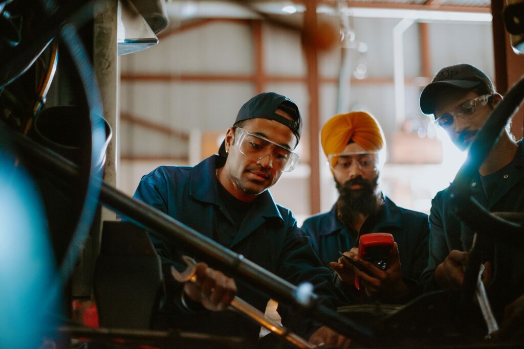 Three student technicians in a workshop troubleshoot machinery, wearing safety glasses and work uniforms. One holds a wrench, another a diagnostic tool.