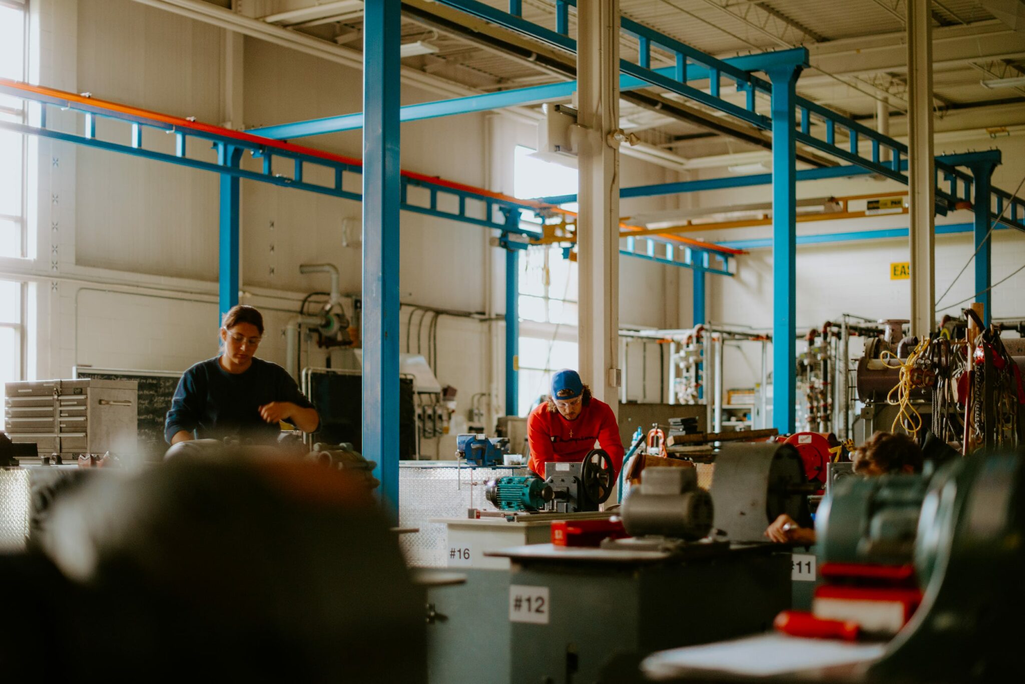 Students in a factory setting operating machinery and equipment amidst metal structures and overhead pipes.