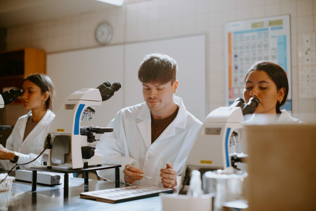 Three student researchers in lab coats use microscopes at a laboratory desk.