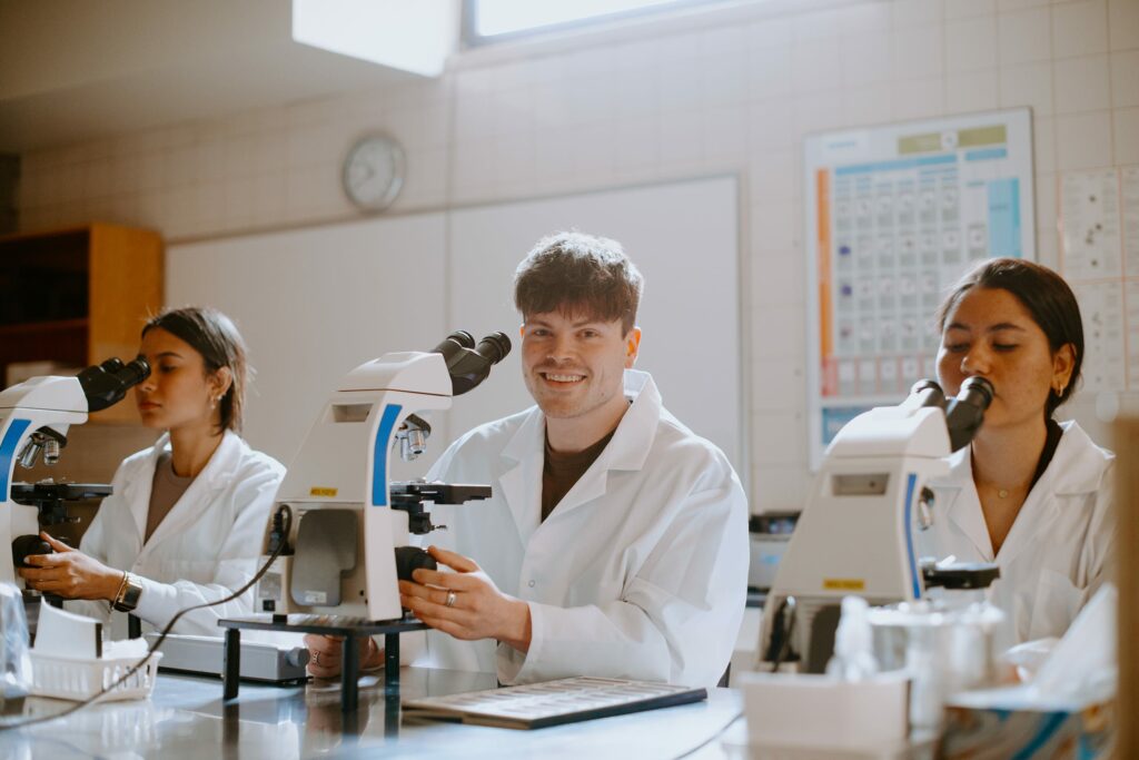 Three student researchers in lab coats use microscopes at a laboratory table, focused on their work.