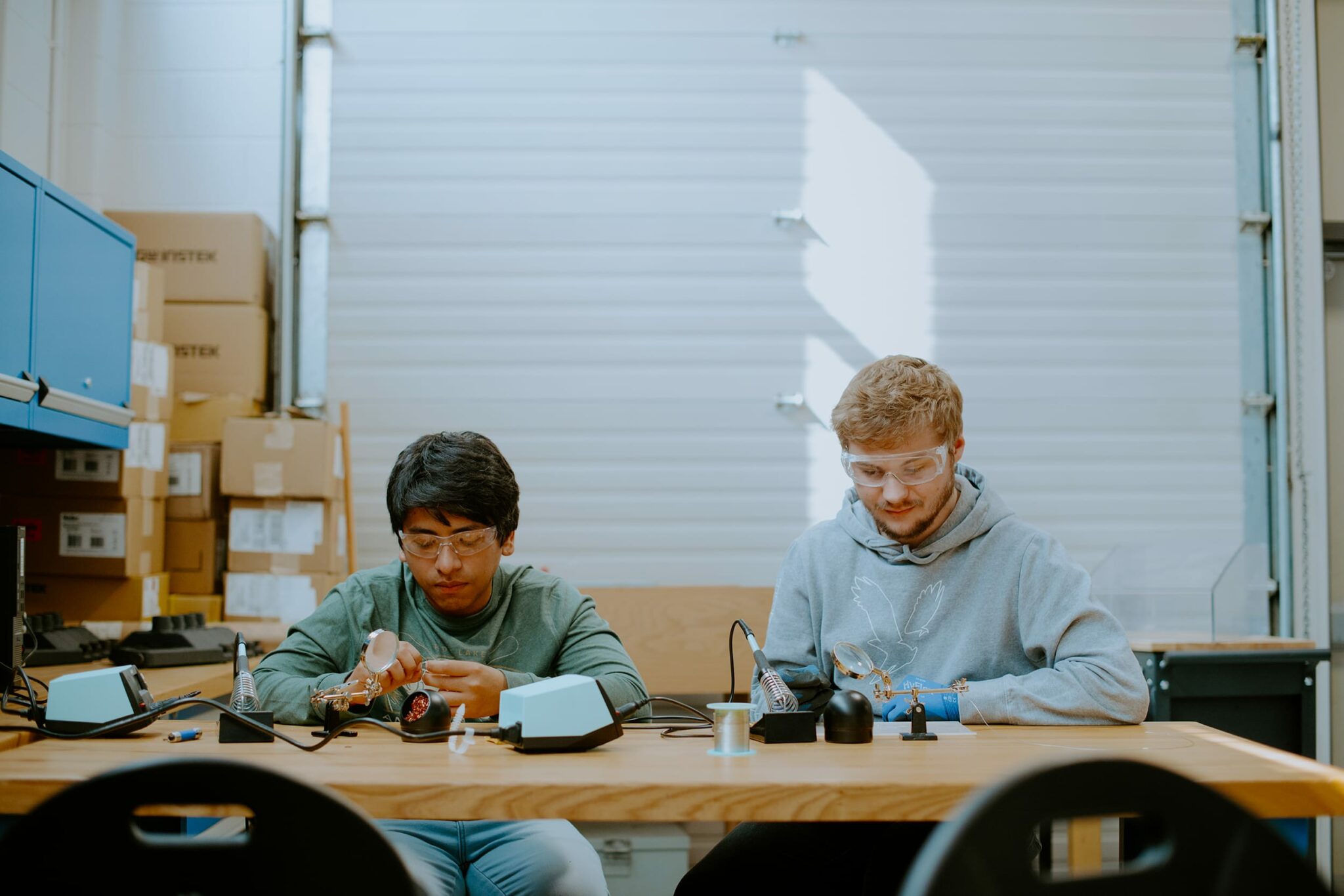 Two students work on electronic circuits at a table, surrounded by soldering equipment and tools in a workshop.