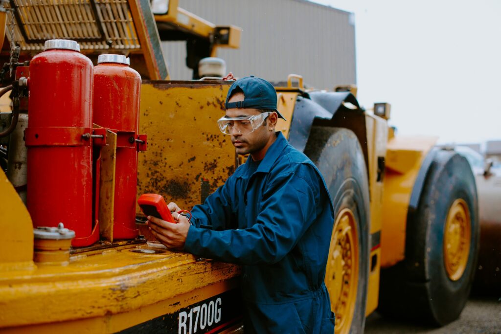A student in blue coveralls and safety gear uses a device near an industrial vehicle with red canisters.
