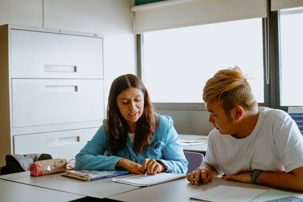 Two students sit at a table in an office, discussing documents on the desk. One is pointing at the paper while the other listens attentively.