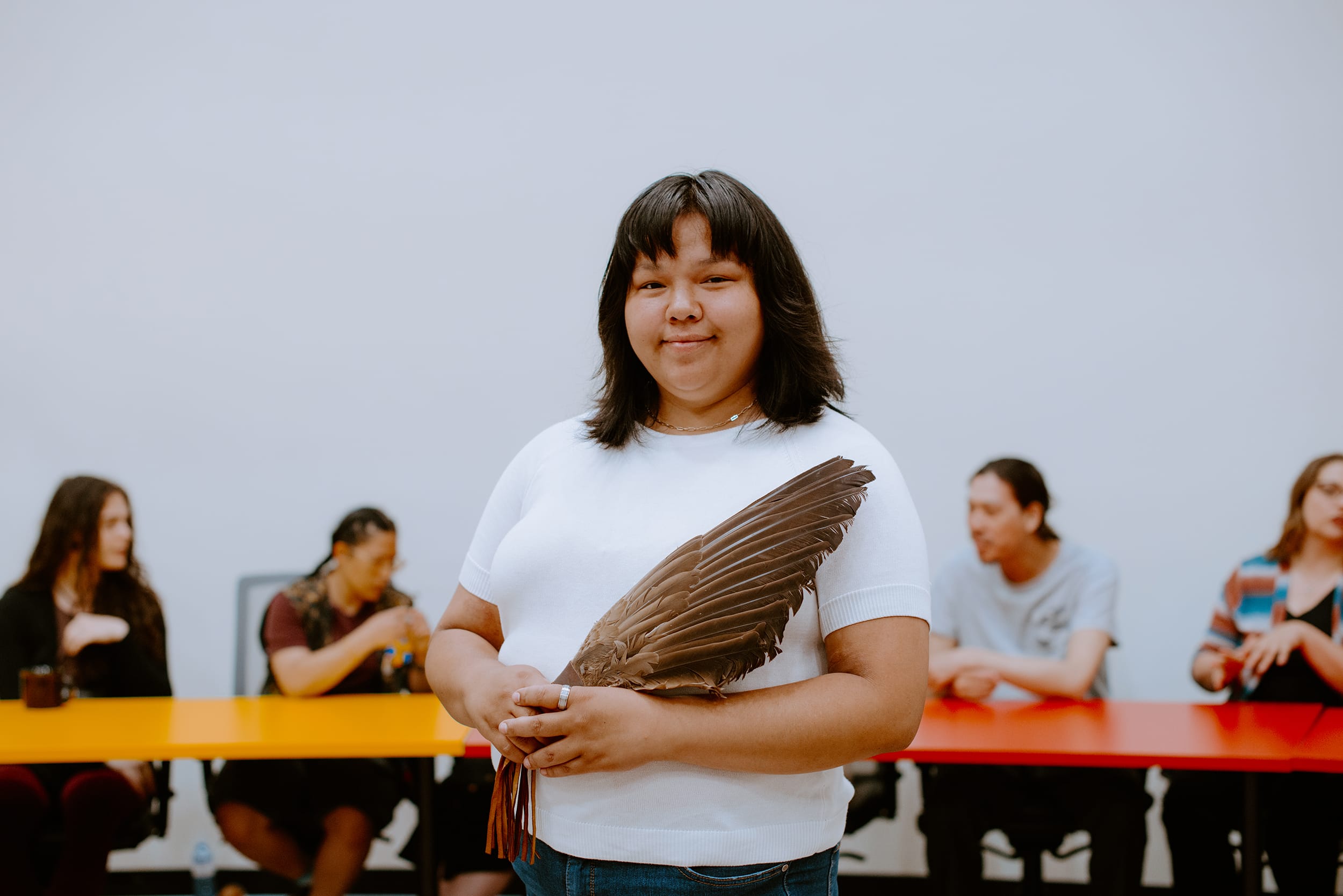 A student is holding feathers stands in front of a group seated at a colorful table.