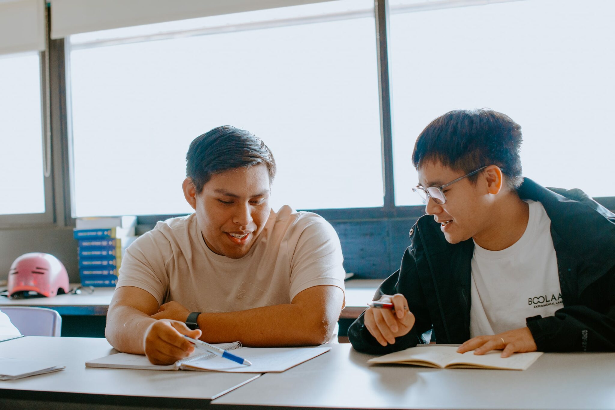 Two students sit at a table in a room with large windows, discussing and writing in notebooks.