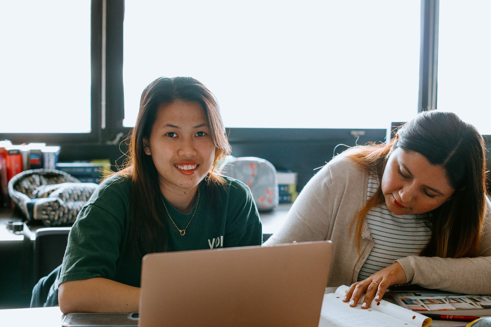 Two students at a desk, one smiling at the camera, the other reading notes. A laptop and books are on the table.