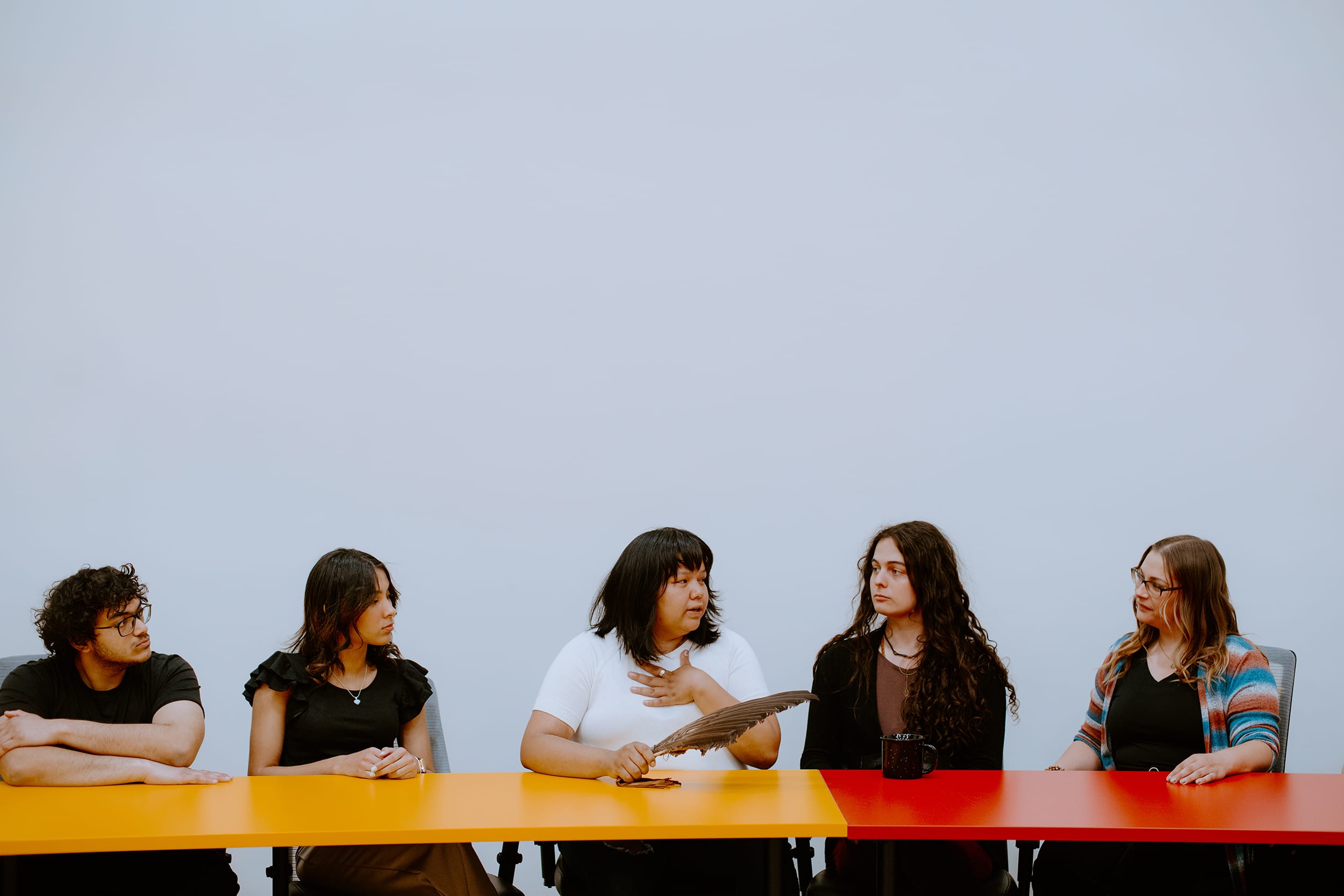 Five students are sitting at a colorful table engaged in discussion. One person is holding a large feather.
