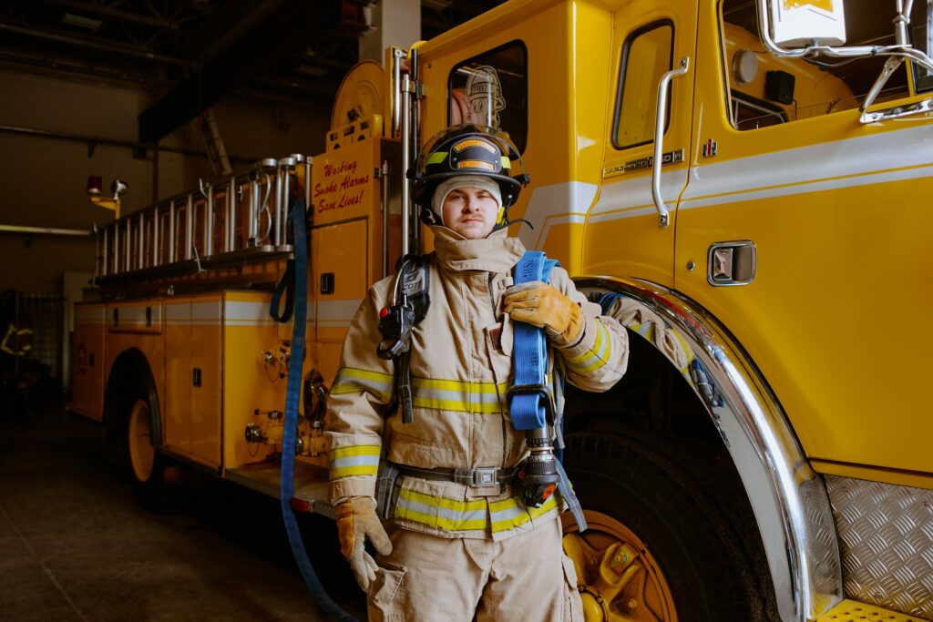 A firefighter student in full gear stands beside a yellow fire truck inside a garage, holding a coiled blue hose.