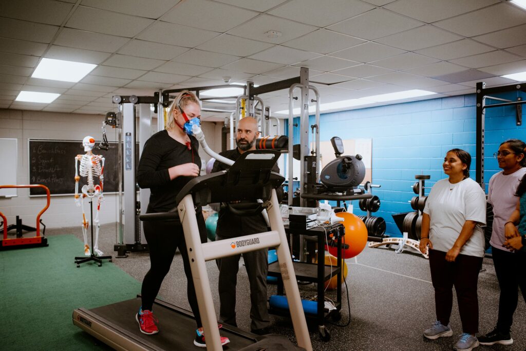 A student exercises on a treadmill with a trainer nearby. Three people observe. The room has gym equipment, including weights and a stationary skeleton model.