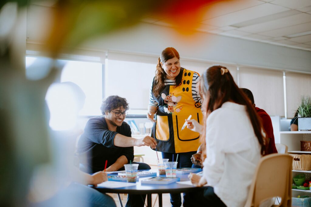 A group of students sit around a table engaged in an activity. A woman wearing a yellow apron stands, smiling, among them.