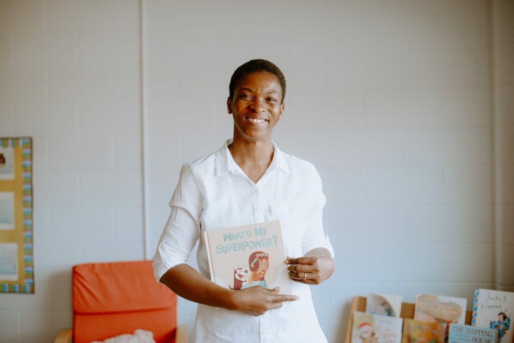 A student in a white shirt smiles while holding a children's book titled "What's My Superpower?" in a room with books and an orange chair.