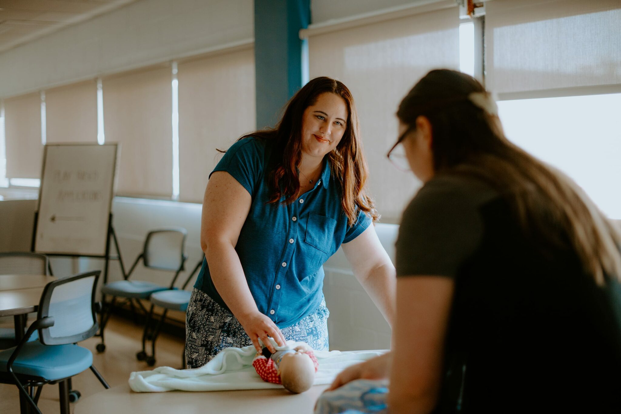 A student demonstrates first aid techniques on a baby mannequin while another person watches in a classroom setting.