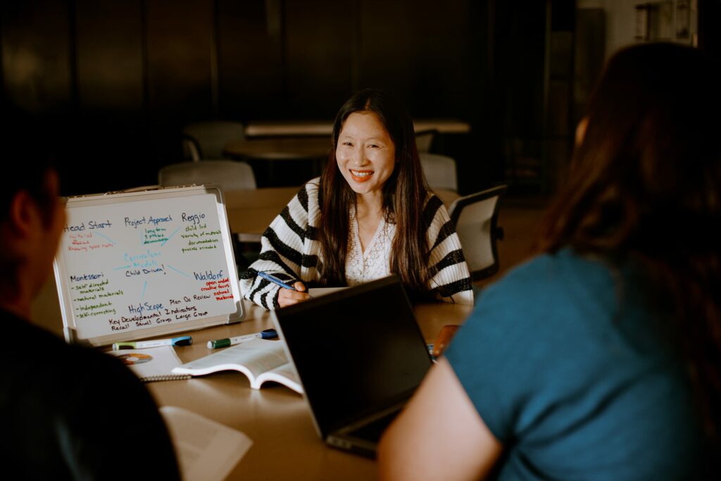 A student is sitting at a table with a laptop and a small whiteboard covered in notes. Two other students are partially visible, focused on the discussion.