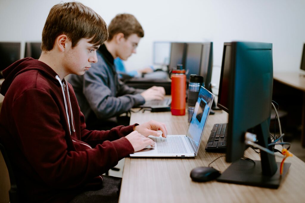 Two students in a classroom work on computers. One of them uses a laptop, while the other uses a desktop. A red water bottle is on the desk.