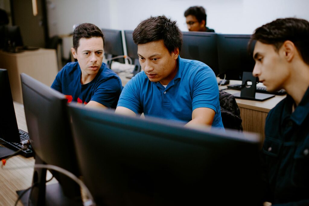 Three students focused on a computer screen in an office setting, discussing work.