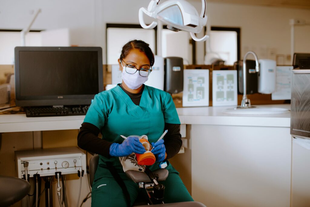 A dental student in a medical setting wears scrubs, mask, and gloves, holding dental tools next to a dental model.