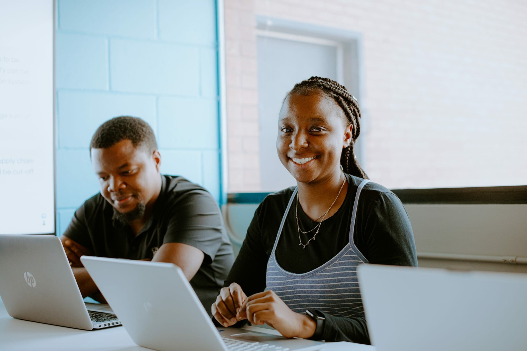 Two students are seated at a table with laptops. They are in a bright room.