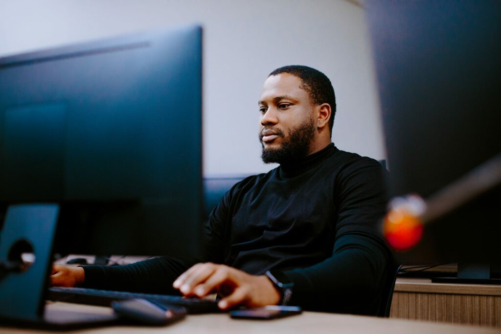 Student working at a computer desk, wearing a black shirt, focused on the screen.