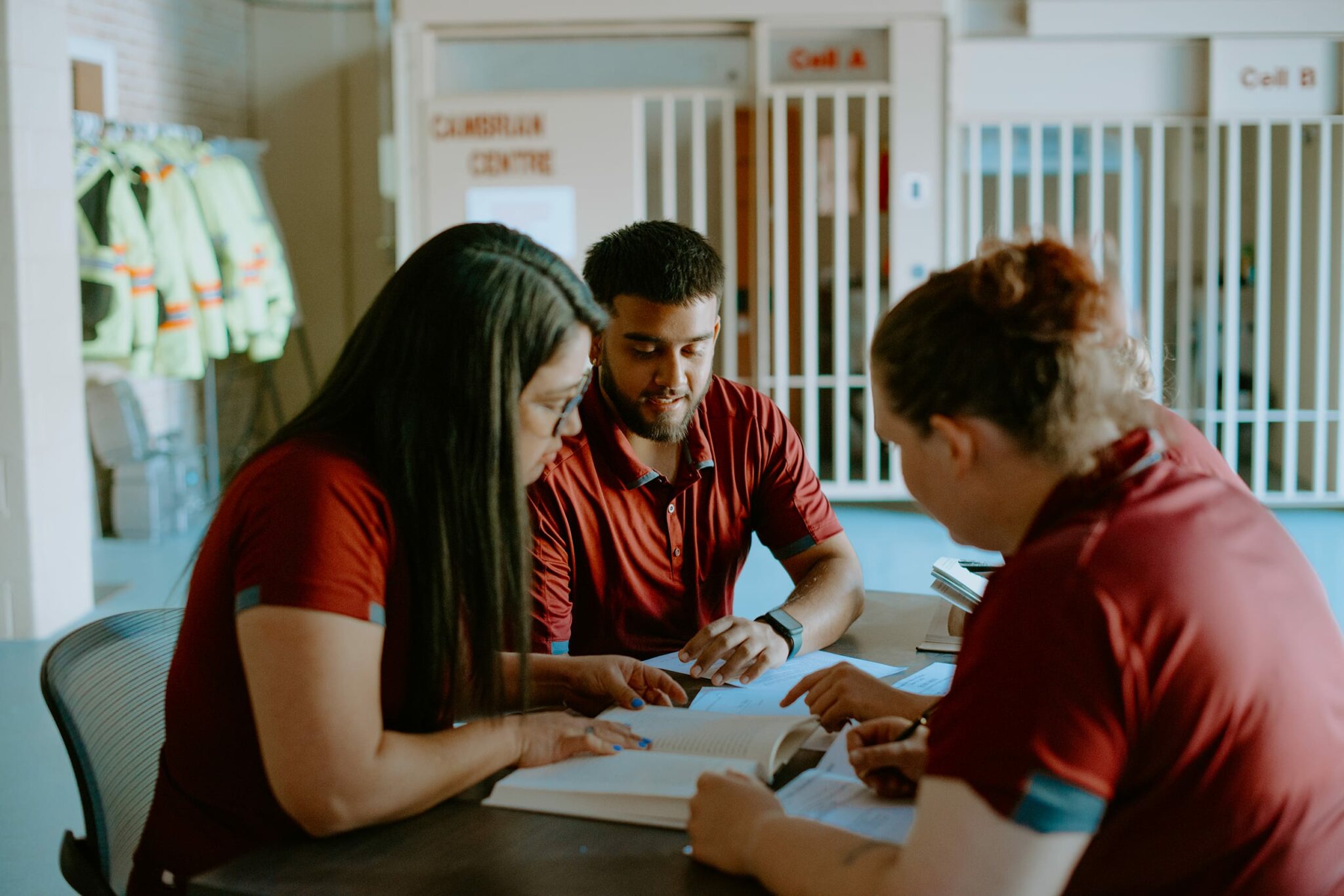 Three students in red shirts sit at a table, engaged in discussion over an open book, in a room with safety gear hanging on the wall.