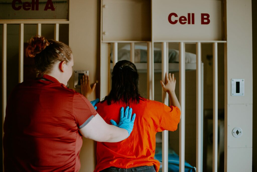 A student in an orange shirt is being patted down by another student in a red shirt and blue gloves, in front of a jail cell marked "Cell B.