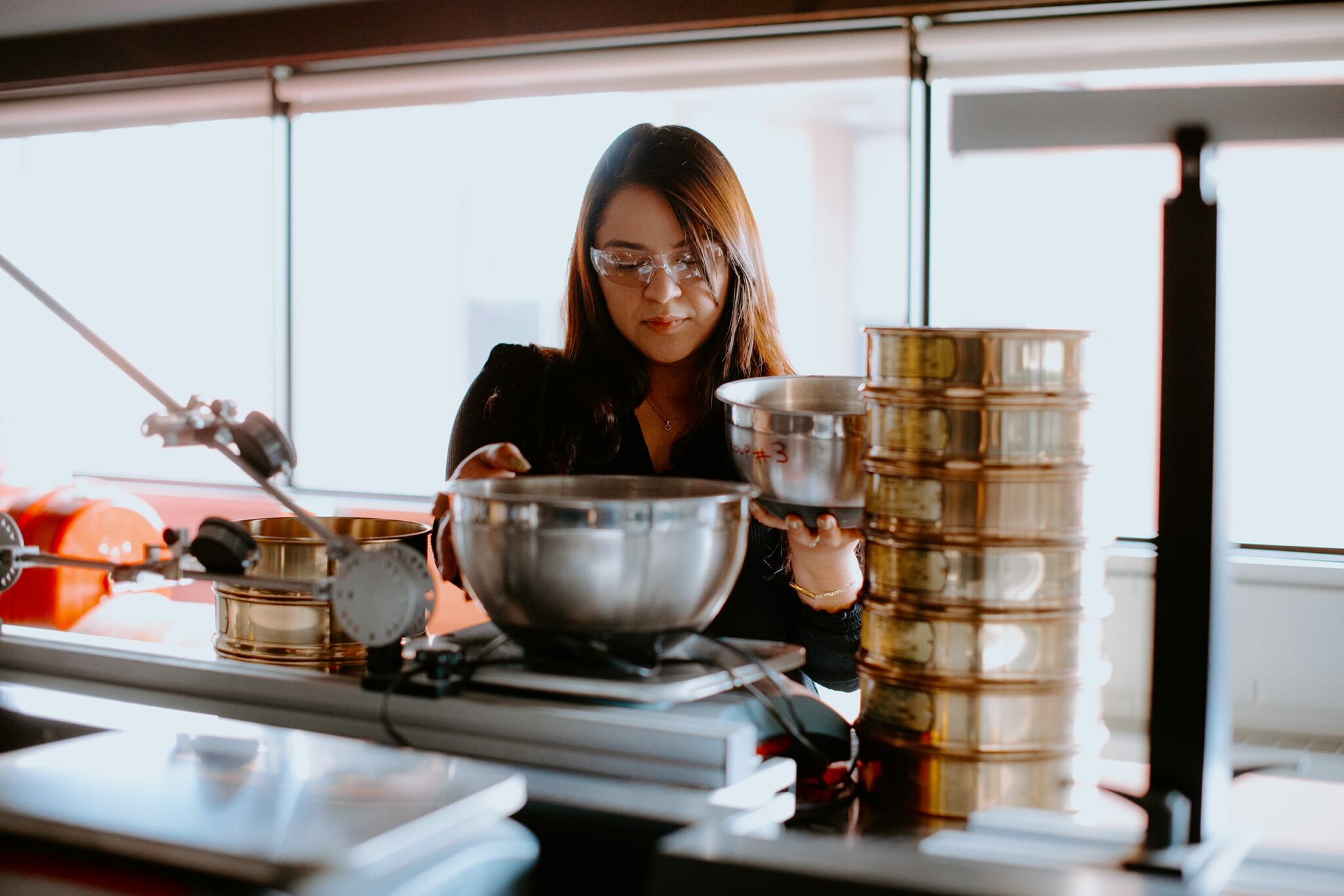A student in safety glasses uses metal equipment and bowls in a laboratory setting.
